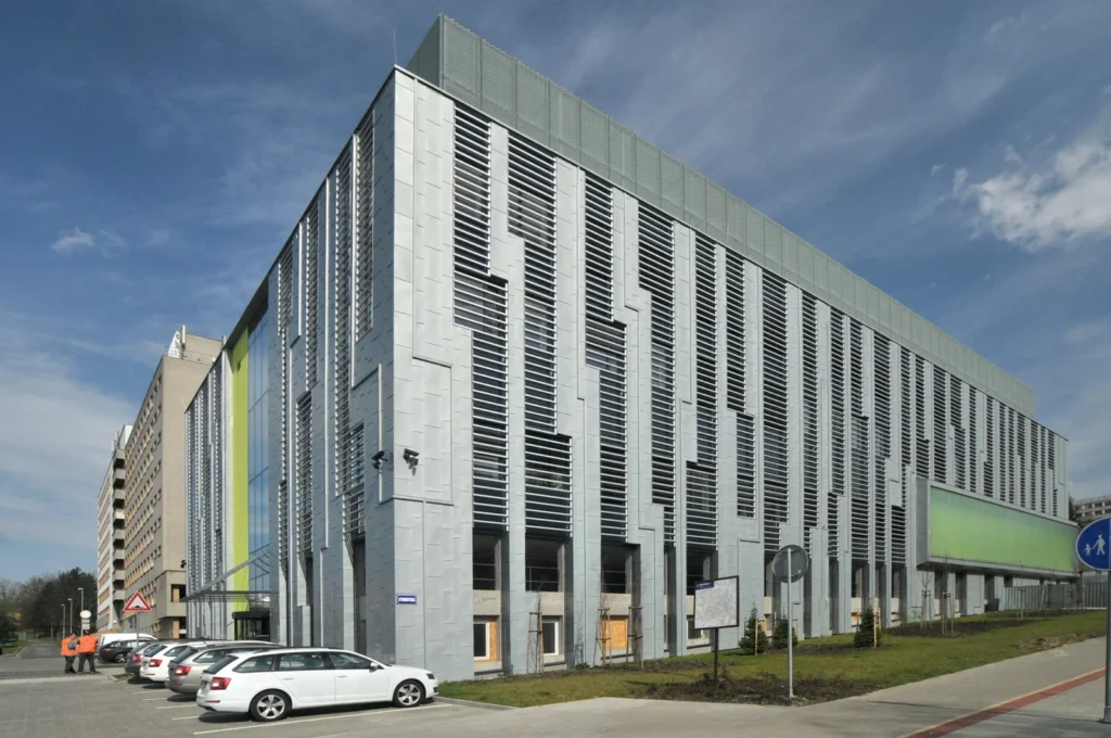 Národní superpočítačové centrum IT4Innovations v Ostravě – moderní architektura a špičkové technologie Budova Národního superpočítačového centra IT4Innovations v Ostravě s moderní kovovou fasádou a výraznými vertikálními prvky, před kterou parkují auta a procházejí lidé.