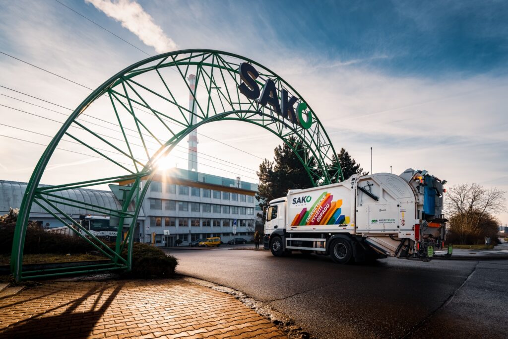 A SAKO Brno waste collection truck drives through the entrance to the company’s site in Brno, with the green SAKO arch and the facility buildings in the background.