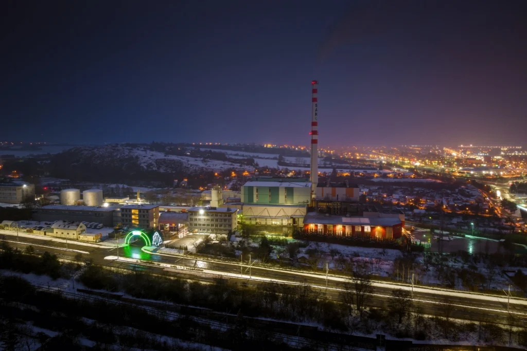 Noční pohled na areál SAKO Brno na Bílé hoře s osvětlenou spalovnou, komínem a nasvíceným vstupním obloukem u hlavní silnice.