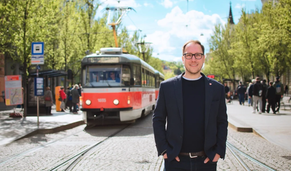 Filip Chvátal stojí na tramvajové trati v Brně, v pozadí projíždí tramvaj a alej stromů.