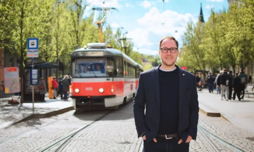 Filip Chvátal stojí na tramvajové trati v Brně, v pozadí projíždí tramvaj a alej stromů.