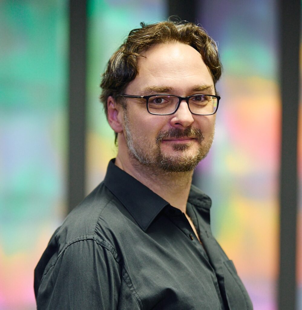 Composer, conductor and violinist Jan Valta in a black shirt, pictured in front of a softly blurred colourful background.